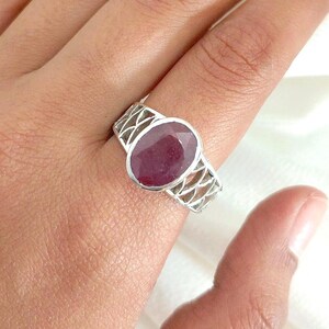 May include: A silver ring with an oval, deep red gemstone. The ring has an openwork design on the band. The ring is shown on a hand against a white background.