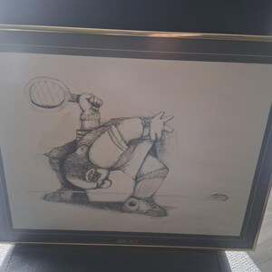 May include: Framed pencil sketch of a tennis player in action, with a racket raised and the ball in motion. The artwork is in a gold-toned frame, depicting a dynamic sports scene. The artist's signature is visible in the bottom right corner.