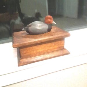 May include: A wooden duck decoy with a gray body, orange head, and white beak rests on a stained wooden box. The box has a rectangular shape with a slightly raised top and bottom. The duck is positioned to the left.