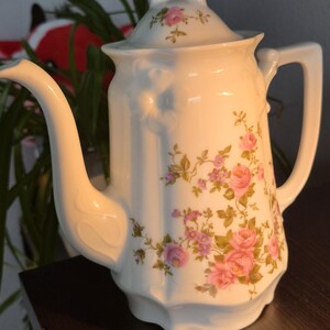 May include: A white porcelain teapot featuring a delicate floral pattern of pink roses and small purple flowers. The teapot has a curved handle, a long spout, and a matching lid with a pointed finial. The body has a subtle vertical ribbed texture.