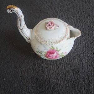 May include: A white ceramic creamer with a curved handle and a spout. The creamer features a pink rose design with gold accents on the handle and rim. A pink rose is on the lid.
