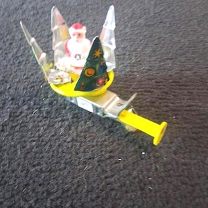 May include: A whimsical Christmas ornament featuring a miniature Santa Claus figure seated in a yellow sleigh. The sleigh is adorned with clear, pointed accents and a small Christmas tree. The ornament has a yellow handle.