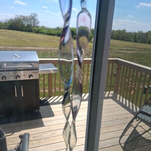 May include: A set of clear, twisted glass wind chimes hanging in front of a window. The chimes have a teardrop shape and reflect the outdoor scenery. The background includes a wooden deck, a grill, and a view of a grassy field under a blue sky.