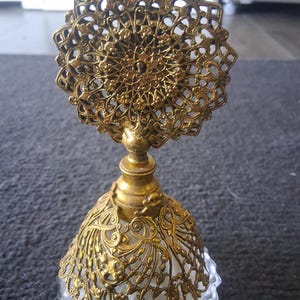 Può includere: Un'antica bottiglia di profumo con una base in vetro trasparente e un rivestimento decorativo in filigrana color oro. La bottiglia presenta una parte superiore ornata a forma di stella e un design floreale dettagliato, che mostra un'estetica vintage.