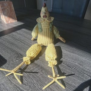 May include: A whimsical, handmade chicken figurine with a yellow body, green overalls, and corn cob legs. The chicken has a red comb and a cream-colored face. The figurine is designed to sit upright, adding a touch of rustic charm to any space.