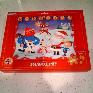 Puede incluir: Caja roja de un rompecabezas familiar de Rudolph, el reno de la nariz roja. La caja presenta una colorida ilustración de Rudolph, Papá Noel y otros personajes del especial navideño. El rompecabezas contiene 530 piezas.