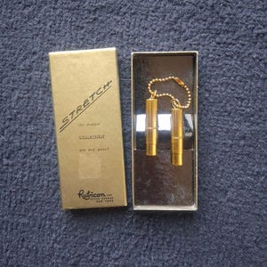 Op de afbeelding: Een goudkleurige pen- en potloodset in een doos. De doos is goudkleurig met het woord "STRETCH" erop gedrukt. De set bevat twee cilindrische pennen die met een ketting zijn verbonden. De doos heeft ook de tekst "Rubicon Inc. Fifth Avenue New York".