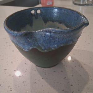 May include: A handmade ceramic bowl with a dark green base and a blue and white glazed rim. The bowl has a pouring spout and two small holes near the rim. The bowl is designed for kitchen use.