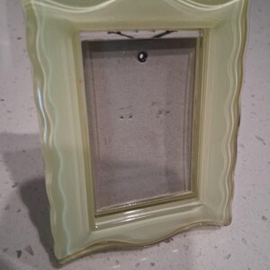 May include: A pale yellow, translucent glass picture frame with a wavy, scalloped edge design. The frame features a rectangular opening with a gray backing board visible inside, including a small circular hole at the top center for hanging.