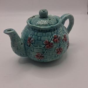 May include: A turquoise ceramic teapot featuring a raised basket-weave texture across the body and lid. The teapot is decorated with small, embossed red clover or shamrock motifs. It includes a curved handle, a spout, and a round lid knob.