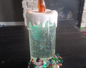 Vela luminosa con efecto de remolino y purpurina, con escena de muñecos de nieve navideños.