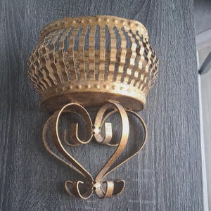 May include: A gold-toned wall sconce with a heart-shaped design. The sconce has a decorative basket-style top with vertical cutouts and a heart-shaped base. The metal has a distressed finish, adding to its vintage appeal.