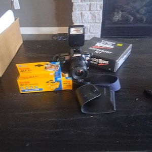 May include: A black Canon EOS Rebel camera with a flash attachment, lens, and a black camera case. Also included are a Kodak Max film box and a Canon EOS Rebel accessory kit box.
