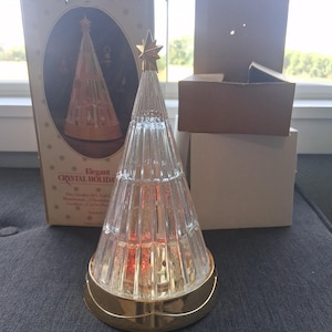 May include: An elegant crystal holiday tree with a gold star topper. The clear glass tree has a gold base and is lit from within. The original box is visible, featuring a similar image of the tree. The tree is approximately 10 inches tall.