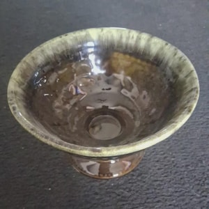 以下が含まれることがあります： 台座付きの茶色のセラミックボウル。ボウルは、幅が広く浅い形状で、内側は濃い茶色、縁は明るいオリーブグリーンです。釉薬は光沢のある仕上がりです。