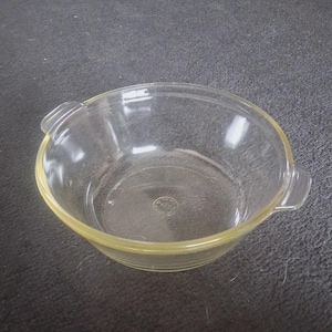 Puede incluir: Un cuenco de vidrio transparente con forma redondeada y dos pequeñas asas. El cuenco tiene un tinte amarillo alrededor del borde. El cuenco es de vidrio Pyrex y está sobre una superficie gris oscuro. Ideal para cocinar.