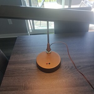 May include: A desk lamp with a wide, rectangular horizontal shade and a circular base in a metallic copper finish. The lamp features a thin vertical stem, a brown power cord, and two small circular buttons, one red and one black, on the base.