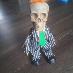 May include: A decorative skull figure with an orange mohawk hairstyle, wearing a black and white striped suit and a green tie. The figure has black shoes and is posed in a seated position. A Halloween-themed decoration.