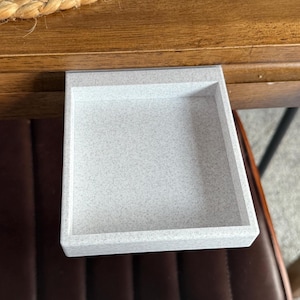 May include: A small, rectangular, light gray tray with a speckled pattern, attached to the underside of a wooden table. The tray is positioned over a brown leather chair, suggesting a storage or organizational function.