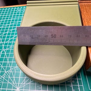 May include: A light green, circular container with a ruler measuring its diameter. The ruler shows a measurement of approximately 10 cm. The container is on a green cutting mat.