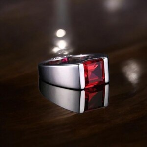 May include: A silver ring with a large, square, ruby-red gemstone. The ring has a polished, reflective surface, and the gemstone is set in a square bezel. The ring is displayed on a dark, reflective surface.