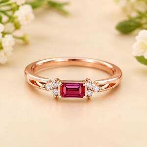 Könnte beinhalten: Ein Ring aus Roségold mit einem zentralen, rechteckigen, im Baguetteschliff geschliffenen, dunkelrosa Edelstein, der auf jeder Seite von drei kleinen, runden, klaren Steinen flankiert wird. Die Ringschiene ist poliert und weist eine geteilte Form auf.