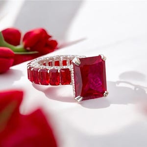 Peut inclure: Une bague en argent ornée d'une grande pierre rouge rectangulaire. La monture est ornée de petites pierres rouges rectangulaires. Des tulipes rouges en arrière-plan créent une composition romantique. La bague est une pièce maîtresse.