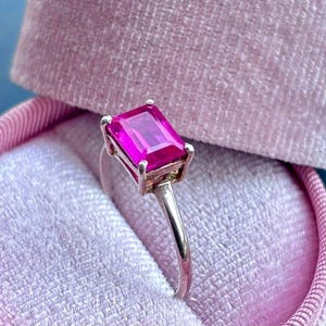 Może przedstawiać: Srebrny pierścionek z prostokątnym, jasno różowym kamieniem szlachetnym. Pierścionek osadzony jest w srebrnej oprawie i prezentowany w różowym aksamitnym pudełku na pierścionek. Kamień szlachetny jest centralnym elementem, który odbija światło i zapewnia żywy kolor.