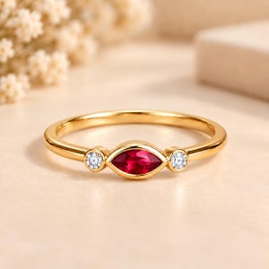 Peut inclure: Une bague en or avec une pierre rouge centrale taille marquise, encadrée par deux petites pierres blanches rondes et transparentes. L'anneau est lisse et poli, avec les trois pierres serties dans des chatons suivant leur forme.