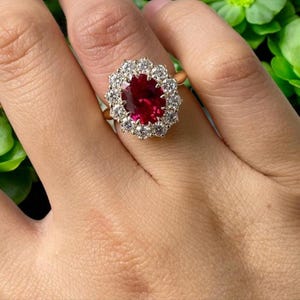 May include: A gold ring featuring a large, oval-cut ruby surrounded by a halo of small, sparkling diamonds. The ring is set on a gold band and is shown on a person's finger. The ruby is a deep red color, and the diamonds are clear and brilliant.