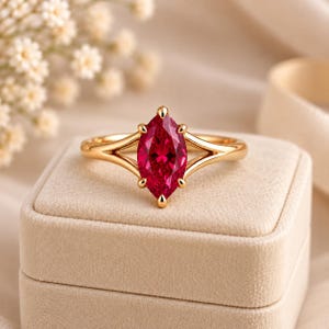 Op de afbeelding: Een gouden ring met een felrode edelsteen in marquise-slijpvorm, vastgehouden door zes pootjes. De ringband heeft een gespleten ontwerp dat taps toeloopt naar de centrale steen. De ring ligt op een vierkant, beige, fluweelachtig doosje.