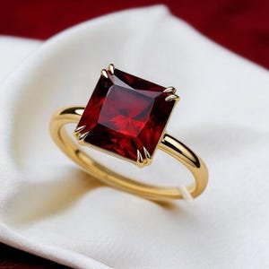 Puede incluir: Un anillo de oro con una gran piedra preciosa roja oscura de talla cuadrada. La piedra preciosa está engastada en una banda de oro con delicadas garras. El anillo se exhibe sobre una tela blanca y sedosa, con un fondo rojo.