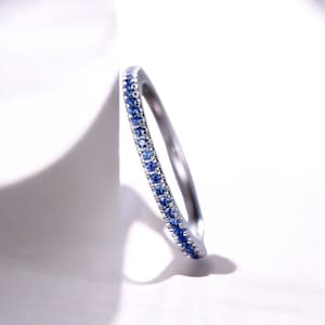 May include: A silver ring with a band of small, round, blue gemstones. The ring is set against a white background, with a shadow cast to the left. The gemstones are a vibrant blue color, and the ring appears to be made of a shiny metal.