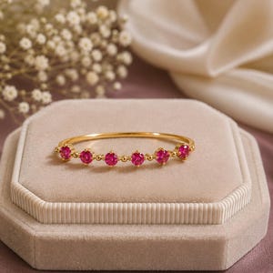 May include: A thin gold band ring featuring five round, vibrant pink gemstones set in a row. Each gemstone is held by small prongs, and the band is accented with tiny gold beads between each stone, creating a delicate and refined appearance.