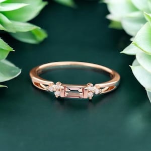 May include: Rose gold ring featuring a rectangular, light pink gemstone at the center, flanked by small, round, clear stones. The ring has a delicate band and is set against a dark green background with blurred green foliage.