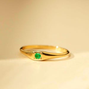 Puede incluir: Un anillo de oro con una pequeña gema verde redonda y brillante en el centro. El anillo tiene un acabado liso y pulido y un diseño simple y elegante. La gema es un contraste vibrante con el oro.
