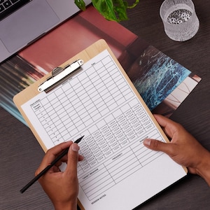 May include: A wooden clipboard with a grid-lined paper, held by hands, with a pen poised to write. A laptop, a plant, and a glass of water are on the dark surface. The paper has columns for frequency and duration.