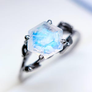 May include: A close-up of a silver ring featuring a large, hexagonal, iridescent gemstone. The gemstone has a blue and white color gradient. The ring's band has a dark, textured design, possibly resembling branches or vines.