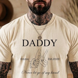 May include: Cream-colored t-shirt with the word "DADDY" in black, featuring a line drawing of hands holding each other. The text "Emma" and "Est.2026" are also present, along with the phrase "Never let go of my hand."
