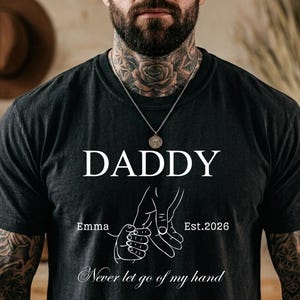 May include: Black t-shirt with the word "DADDY" in white, a graphic of two hands holding each other, the name "Emma", and the year "Est.2026". The phrase "Never let go of my hand" is written at the bottom.
