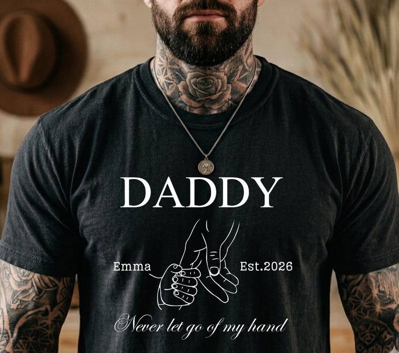 May include: Black t-shirt with the word "DADDY" in white, a graphic of two hands holding each other, the name "Emma", and the year "Est.2026". The phrase "Never let go of my hand" is written at the bottom.