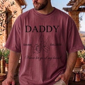 May include: A maroon t-shirt with the word "DADDY" in large black letters. Below the word are two hands holding each other, with the text "Emma" and "Est. 2026". The phrase "Never let go of my hand" is written below the hands.