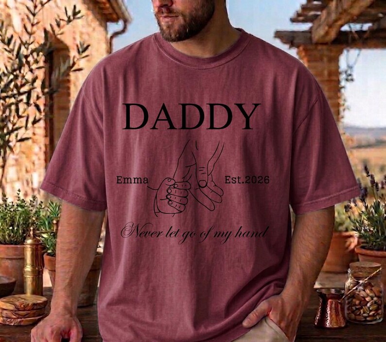 May include: A maroon t-shirt with the word "DADDY" in large black letters. Below the word are two hands holding each other, with the text "Emma" and "Est. 2026". The phrase "Never let go of my hand" is written below the hands.
