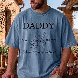 May include: A light blue t-shirt with the word "DADDY" in black, above a line drawing of two hands holding each other. The text "Emma Est. 2026" and "Never let go of my hand" are also printed on the shirt.
