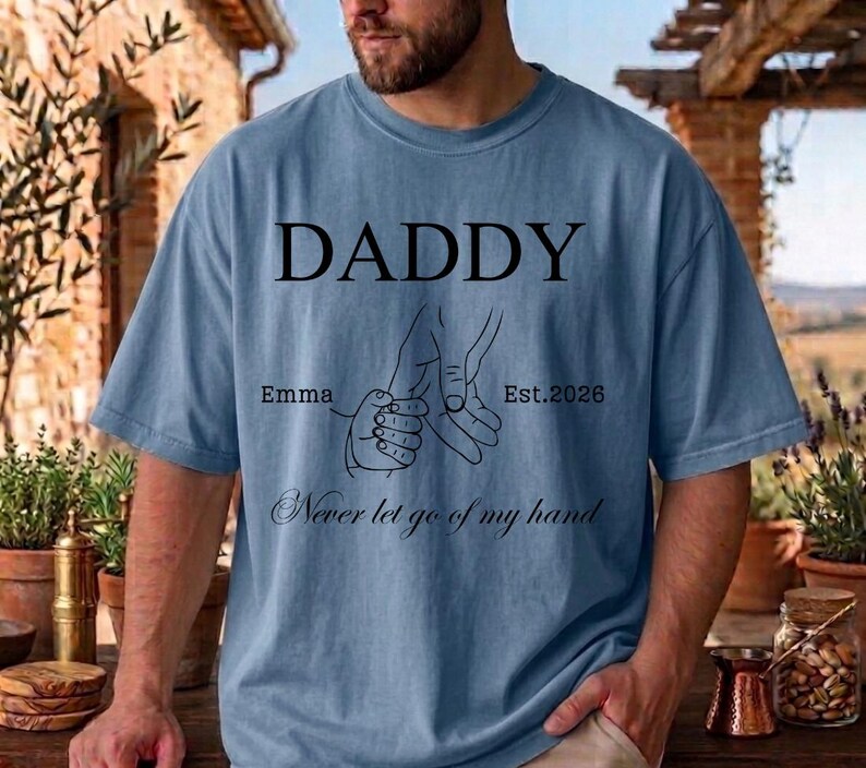 May include: A light blue t-shirt with the word "DADDY" in black, above a line drawing of two hands holding each other. The text "Emma Est. 2026" and "Never let go of my hand" are also printed on the shirt.