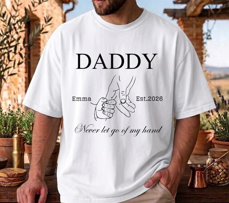 May include: White t-shirt with the word "DADDY" in large black letters. Below the word are line drawings of two hands holding each other, with the text "Emma Est.2026" and "Never let go of my hand" in script.