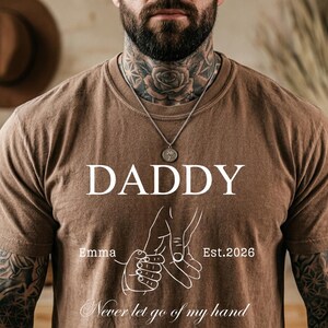 May include: Brown t-shirt with the word "DADDY" in white block letters. Below the word "DADDY" is a graphic of two hands holding each other. The text "Emma" and "Est.2026" are also on the shirt. The phrase "Never let go of my hand" is at the bottom.