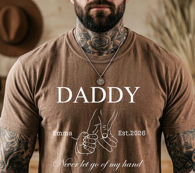May include: Brown t-shirt with the word "DADDY" in white block letters. Below the word "DADDY" is a graphic of two hands holding each other. The text "Emma" and "Est.2026" are also on the shirt. The phrase "Never let go of my hand" is at the bottom.