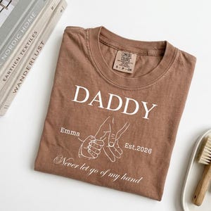 May include: A brown t-shirt with the word "DADDY" in white, along with the name "Emma" and the year "Est.2026". The shirt also features a line drawing of two hands holding each other, with the phrase "Never let go of my hand".