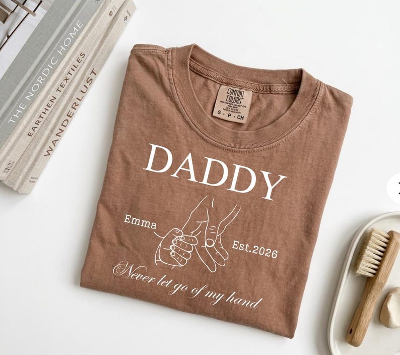 May include: A brown t-shirt with the word "DADDY" in white, along with the name "Emma" and the year "Est.2026". The shirt also features a line drawing of two hands holding each other, with the phrase "Never let go of my hand".